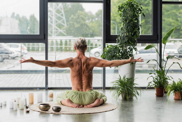 back view of shirtless man sitting in thunderbolt yoga pose near candles and Tibetan singing bowls in studio  - Foto, imagen
