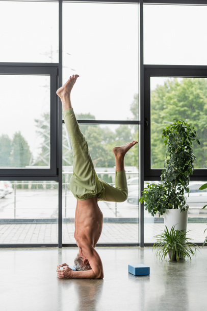 grijs harige man doet ondersteunde headstand in de buurt van yoga schuim blok  - Foto, afbeelding