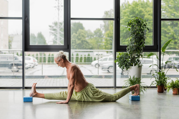 side view of shirtless and grey haired man doing split on yoga foam blocks  - Zdjęcie, obraz
