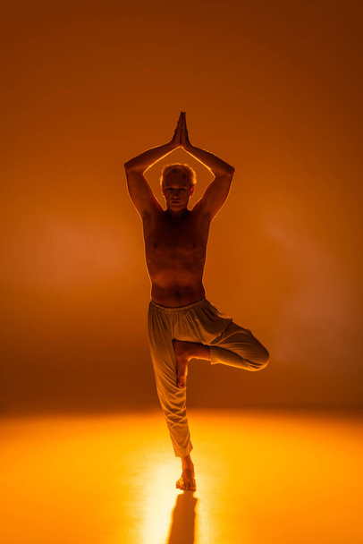 full length of shirtless man in pants standing in tree pose on orange background  - Фото, изображение