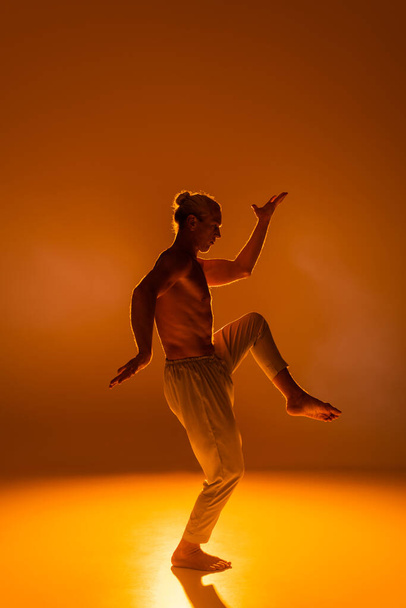 side view of shirtless man in pants practicing yoga on orange background  - Foto, Imagen
