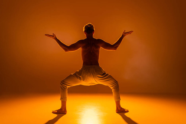 back view of shirtless man in pants practicing goddess yoga pose on orange background  - Foto, imagen