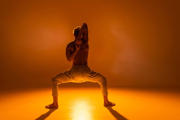 back view of shirtless man practicing goddess yoga pose with clenched hands behind back on orange background  - Photo, image