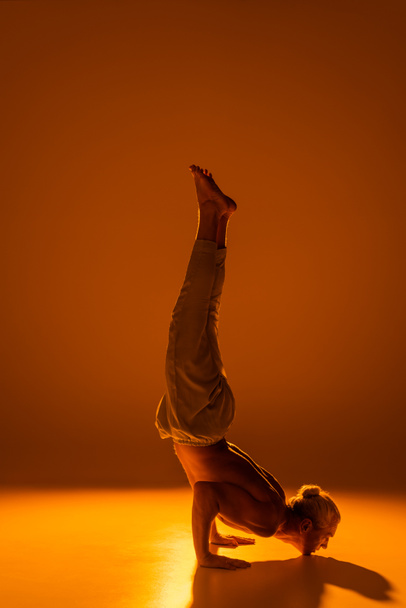 full length of shirtless man in pants doing chin stand yoga pose on brown  - Foto, Bild