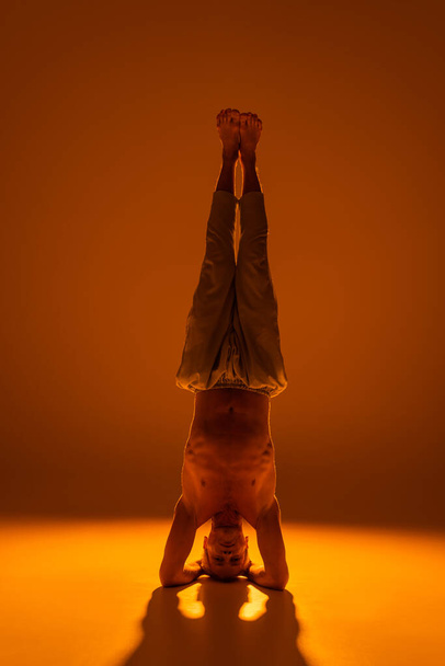 volledige lengte van shirtless man doen ondersteunde headstand yoga pose op bruin  - Foto, afbeelding