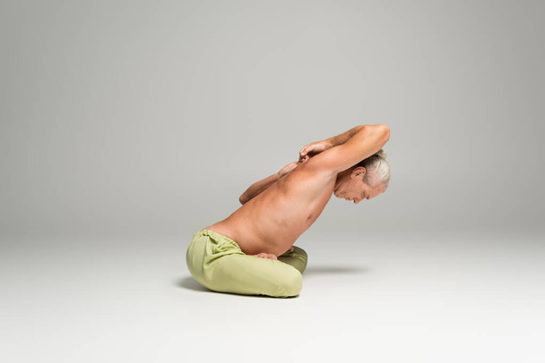 side view of shirtless man sitting in lotus pose with clenched hands behind back on grey background  - Fotografie, Obrázek