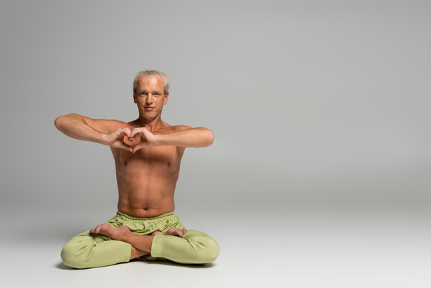 shirtless man in green pants sitting in lotus pose and showing heart sign on grey background  - Foto, imagen