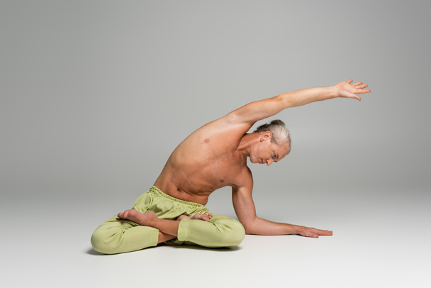 shirtless man in green pants sitting in lotus pose and doing side bend on grey background  - Photo, Image
