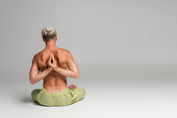 back view of shirtless man in green pants sitting in lotus pose and doing anjali mudra behind back on grey background  - Foto, immagini