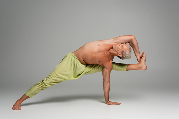 barefoot man in pants doing compass yoga pose on grey background  - Foto, Imagen