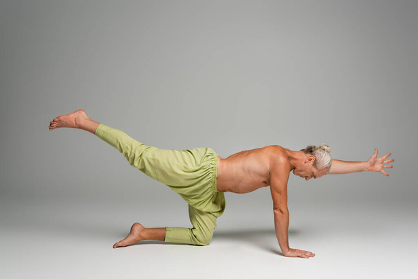 Pantolonlu çıplak ayaklı bir adam, gri üzerinde kaplan yogası pozu veriyor.  - Fotoğraf, Görsel