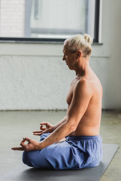 side view of grey haired man in blue pants sitting in lotus pose and doing gyan mudra - Foto, Imagen