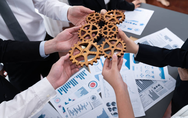 Closeup top view businesspeople hand holding gear and join together over meeting table with financial report papers. Cohesive group of office workers holding cog wheel as synergy harmony concept. - Foto, imagen