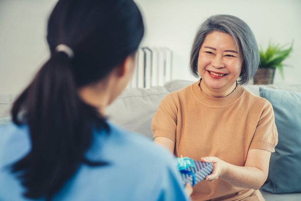 A young caregiver hand over to her senior patient a blue gift box with blue ribbons at a contented living room. - Photo, image