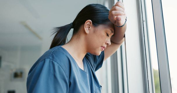 Healthcare nurse, stress and headache by window for working burnout, frustrated employee and tired asian woman in hospital. Doctor, pain and sad medical worker, anxiety or mental health depression. - Photo, Image