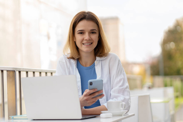 Confident smiling young woman holding mobile phone, using laptop computer looking at camera, communication online sitting in modern street cafe. Technology concept - Photo, Image