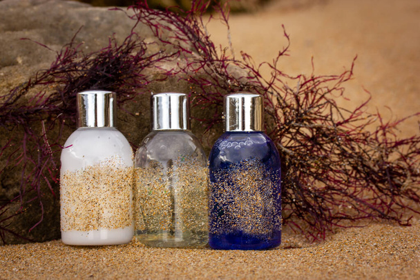 Plastic flessen, containers met eco cosmetica - shampoo, douchegel, bodylotion voor gezichtsverzorging - aan de zandkust tussen het rode zeewier. Natuurlijke cosmetica voor een vakantie aan zee in de zomer - Foto, afbeelding