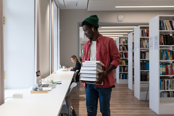 Бібліотеки та онлайн-навчання. Афроамериканець, який тримає купу книжок і дивиться на екран ноутбука під час навчання в бібліотеці університету. Навчання і концепція освіти - Фото, зображення
