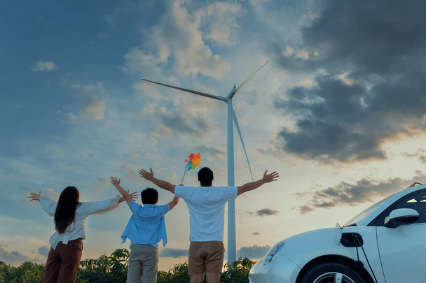 Fogalma progresszív boldog család élvezik idejüket szélerőműben elektromos jármű. Szélturbina-generátorból származó tiszta megújuló energiával hajtott elektromos jármű töltőállomáshoz. - Fotó, kép
