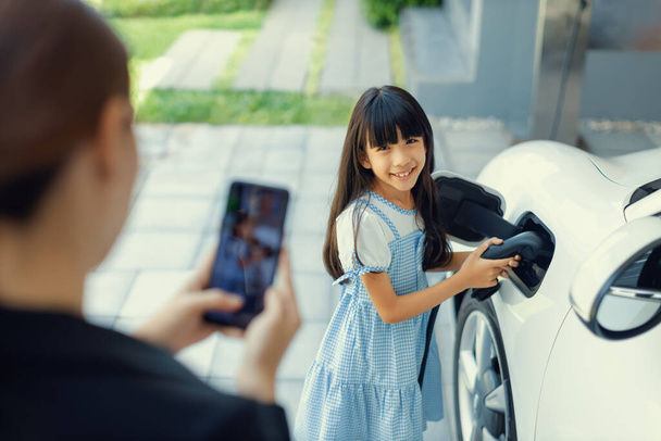 Progressive mother using smartphone take focus photo of daughter recharge EV car. Image of asian girl insert electric charger from home charging station to EV car on phone screen with blur background. - Foto, Imagem