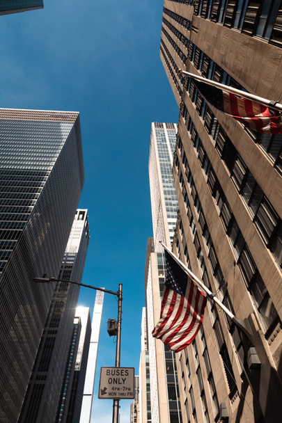nízký úhel pohledu na budovy s vlajkami USA proti modré obloze v okrese Manhattan v New Yorku - Fotografie, Obrázek