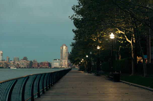 valoisat lyhdyt lähellä puita Hudson joen rantakadulla ja Manhattanin iltakaupunkikuvaa New Yorkissa - Valokuva, kuva