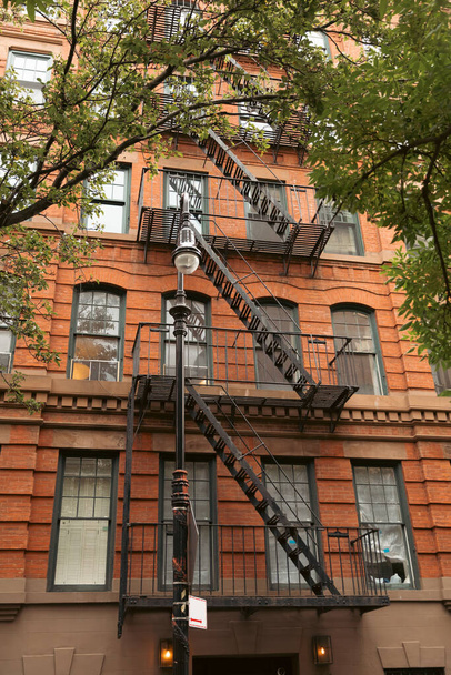 brick dwelling building with metal balconies and fire escape stairs near lantern and trees in New York City - Photo, Image