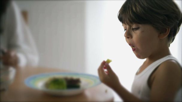 Little boy eating broccoli at lunch. Child sitting at meal table feeding super food vegetable - 写真・画像