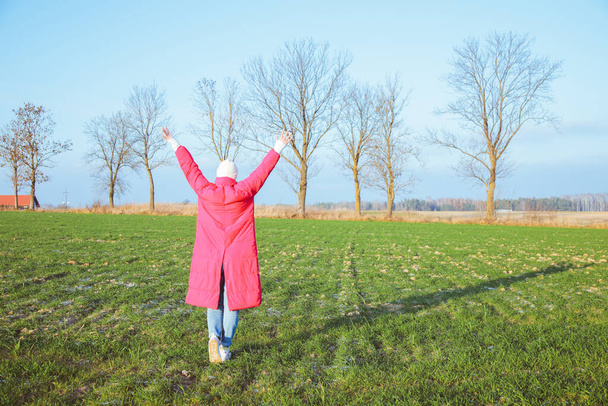 Woman in bright pink coat stand turning away with raised arms in open field. Female full length photo, back view. Green grass, bare trees, blue sky. Picturesque autumn nature. Freedom. - Foto, Imagem
