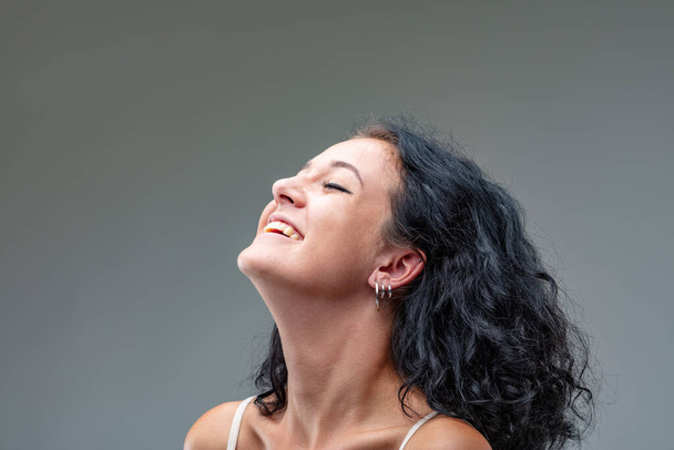 Portrait of young brunette woman in three-quarters . She has closed eyes, bare shoulders and white tank top straps. She laughs happily as she lifts her chin. Her skin is smooth and a healthy color bec - Foto, Bild