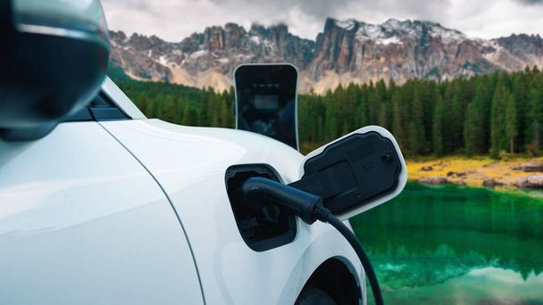 Ενέργεια βιώσιμη δύναμη αυτοκινήτων με ηλεκτρογεννήτρια κίνησης, επαναφόρτιση της μπαταρίας στο σταθμό φόρτισης με φόντο βουνό για προοδευτική ταξιδιωτική έννοια. EV αυτοκίνητο στη φύση ως σύμβολο για καθαρό περιβάλλον. - Φωτογραφία, εικόνα