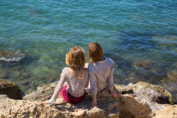 two adult girls sit with their backs on the rocks against the backdrop of the turquoise sea. Mom and adult daughter, girlfriends, sisters on vacation, enjoy the moment - Foto, Imagen