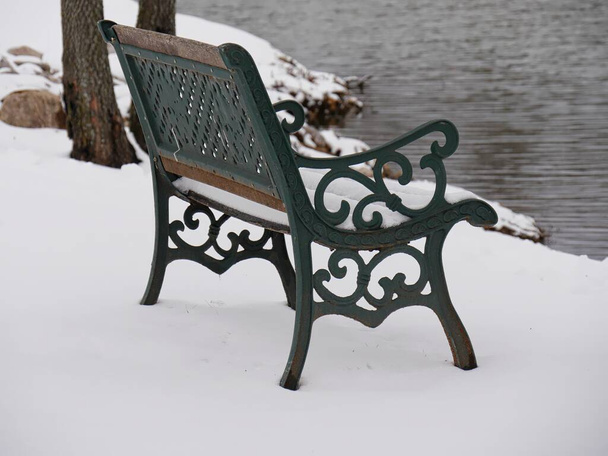 Wrought-iron bench facing the lake on a snow-covered ground - Фото, изображение