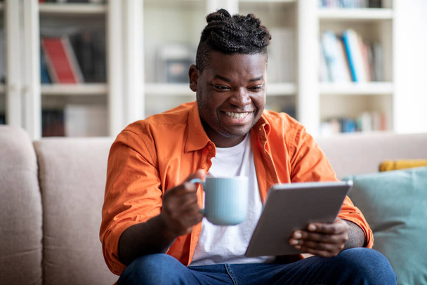 Happy smiling relaxed young chubby african american man in casual outfit with braids sitting on couch at home, using modern digital tablet, watching video content and drinking coffee, copy space - Photo, image