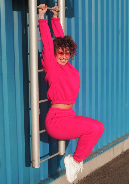 Happy young woman with curly hair in magenta suit and sunglasses posing on blue background. - Foto, Imagem