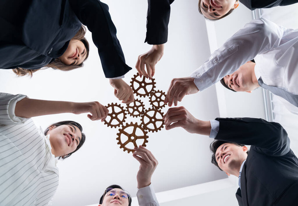 Hand holding wooden gear by businesspeople wearing suit for harmony synergy in office workplace. Bottom view office worker hand make chain of gear into collective unity form. - Valokuva, kuva
