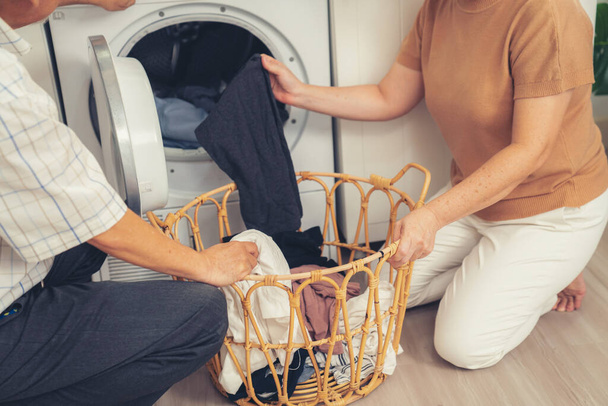 Ηλικιωμένο ζευγάρι που εργάζονται μαζί για να ολοκληρώσουν τις δουλειές του σπιτιού τους στο πλυντήριο ρούχων με έναν ευτυχισμένο και ικανοποιημένο τρόπο. Σύζυγος και σύζυγος κάνουν τις συνήθεις εργασίες στο σπίτι. - Φωτογραφία, εικόνα