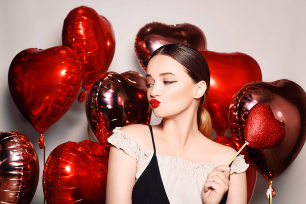 Kaunis tyttö, malli ilmapallojen taustalla sydämen muotoisena. Nainen, jolla on punaiset huulet meikki, ilmapusu ja hyvällä tuulella, rakkaus, tunteet. Ystävänpäivä ja 8 maaliskuu - Valokuva, kuva