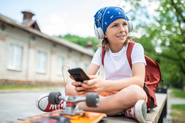 Cute kid in the wireless headphones and with the backpack over his shoulders sitting cross-legged on a wooden bench - Фото, изображение