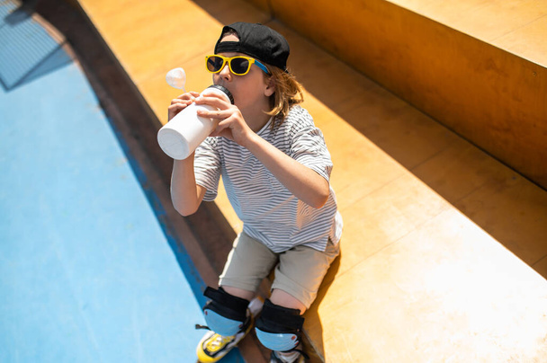 Хлопчик на подушечках і сонцезахисних окулярах сидить на лавці і п'є воду з пластикової спортивної пляшки
 - Фото, зображення