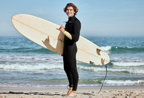 Man, surfer and surfboard at the beach for training exercise or fitness workout in the outdoors. Portrait of happy sports professional with smile showing hand gesture for hang loose ready for surfing. - Fotoğraf, Görsel