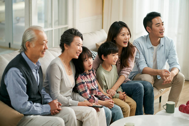 Üç kuşak Asyalı aile evde kanepede oturup birlikte televizyon seyrediyor. Mutlu ve gülümsüyor. - Fotoğraf, Görsel