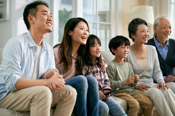 trzy pokolenia azjatycka rodzina siedzi na kanapie w domu oglądając telewizję razem szczęśliwy i uśmiechnięty - Zdjęcie, obraz