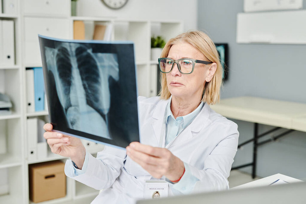 Ernstige volwassen radioloog in bril kijken naar röntgenfoto van de longen van de patiënt tijdens het zitten door de werkplek in het medisch kantoor - Foto, afbeelding