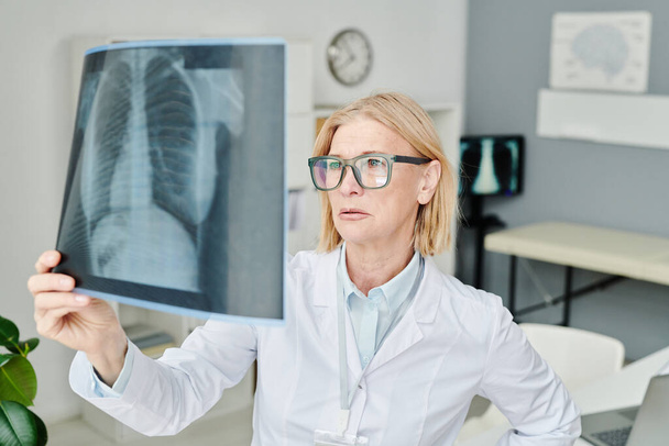 Зрелый опытный рентгенолог в белой юбке, изучающий ронтген легких при попытке установить точный диагноз пациента - Фото, изображение