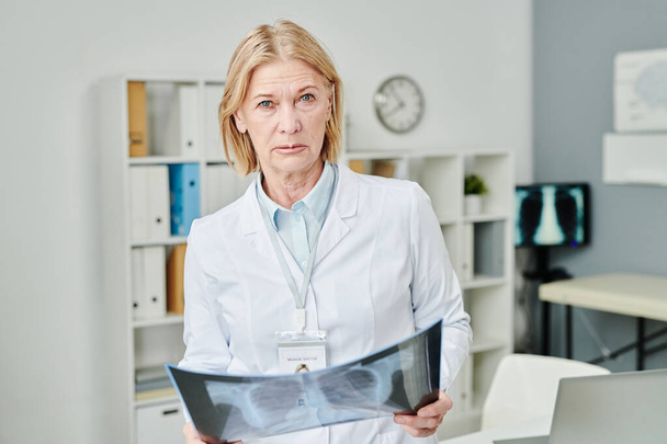 Серьёзная зрелая женщина-врач с рентгеновским снимком легких пациента, смотрящая в камеру, стоя напротив полок и компьютера в медицинском кабинете - Фото, изображение