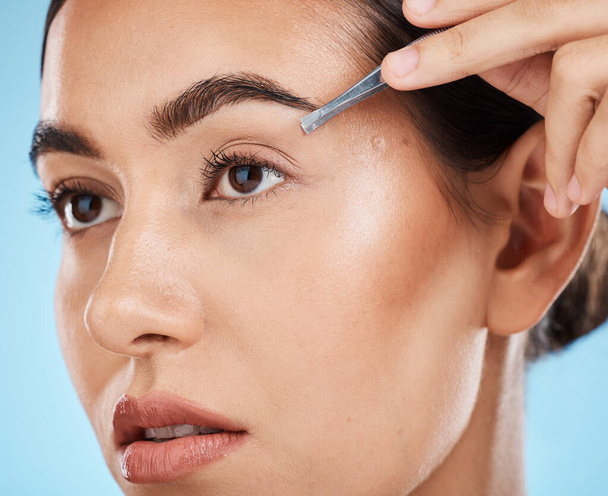 Hair removal, tweezer and woman doing her eyebrows in studio for beauty, facial and grooming routine. Cosmetics, depilation and female model plucking face hair with cosmetic tool by blue background - Fotoğraf, Görsel
