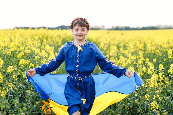 Pray for Ukraine. Child with Ukrainian flag in rapeseed field. A girl in an embroidered shirt runs across the field with the Ukrainian flag in her hands - Foto, immagini