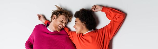 top view of happy redhead man and curly african american woman in magenta color clothes lying on white, banner  - Фото, изображение