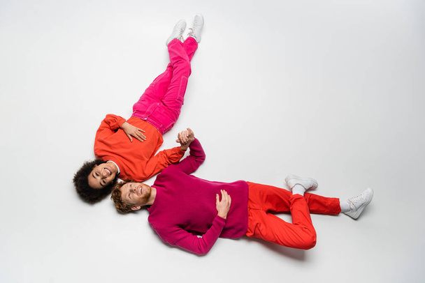 top view of joyful redhead man and curly african american woman in magenta color clothes lying on white  - 写真・画像